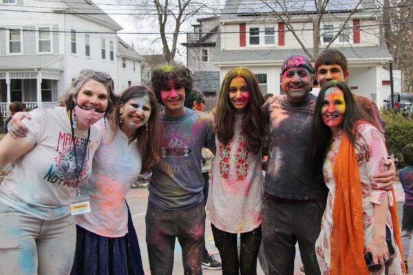 Seven people with colorful powder all over their faces and clothes smile at the camera