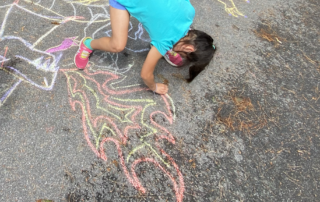 A child uses chalk to draw a dragon on the driveway of the New Art Center during summer art camp.