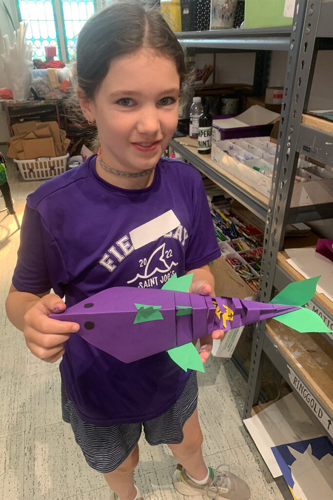 A student in a purple t-shirt holds a purple fish constructed from paper