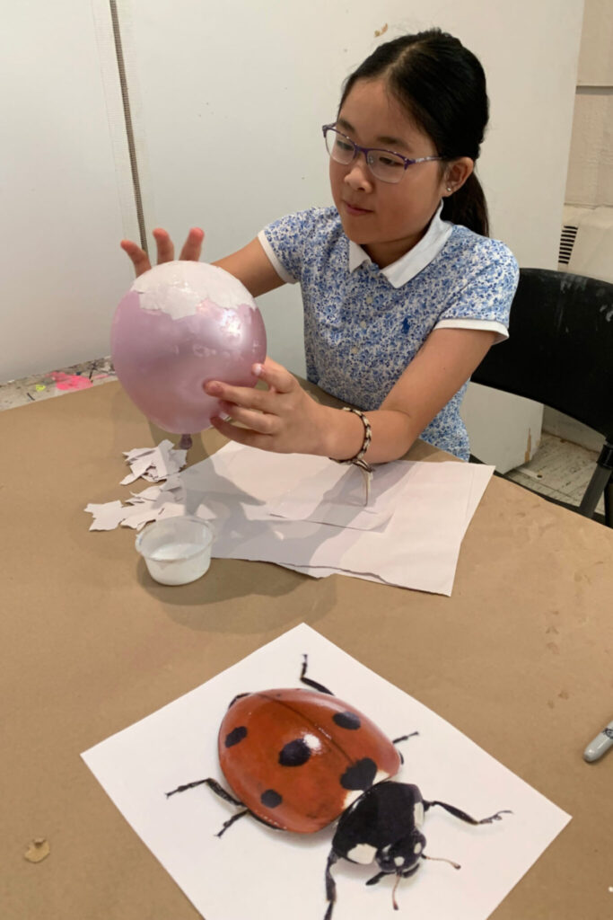 A child uses bits of paper and a glue solution to coat the outside of a balloon.