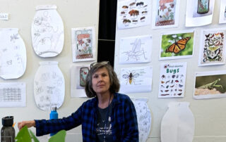 A teacher sits in front of a white board that has many images and drawings of bugs