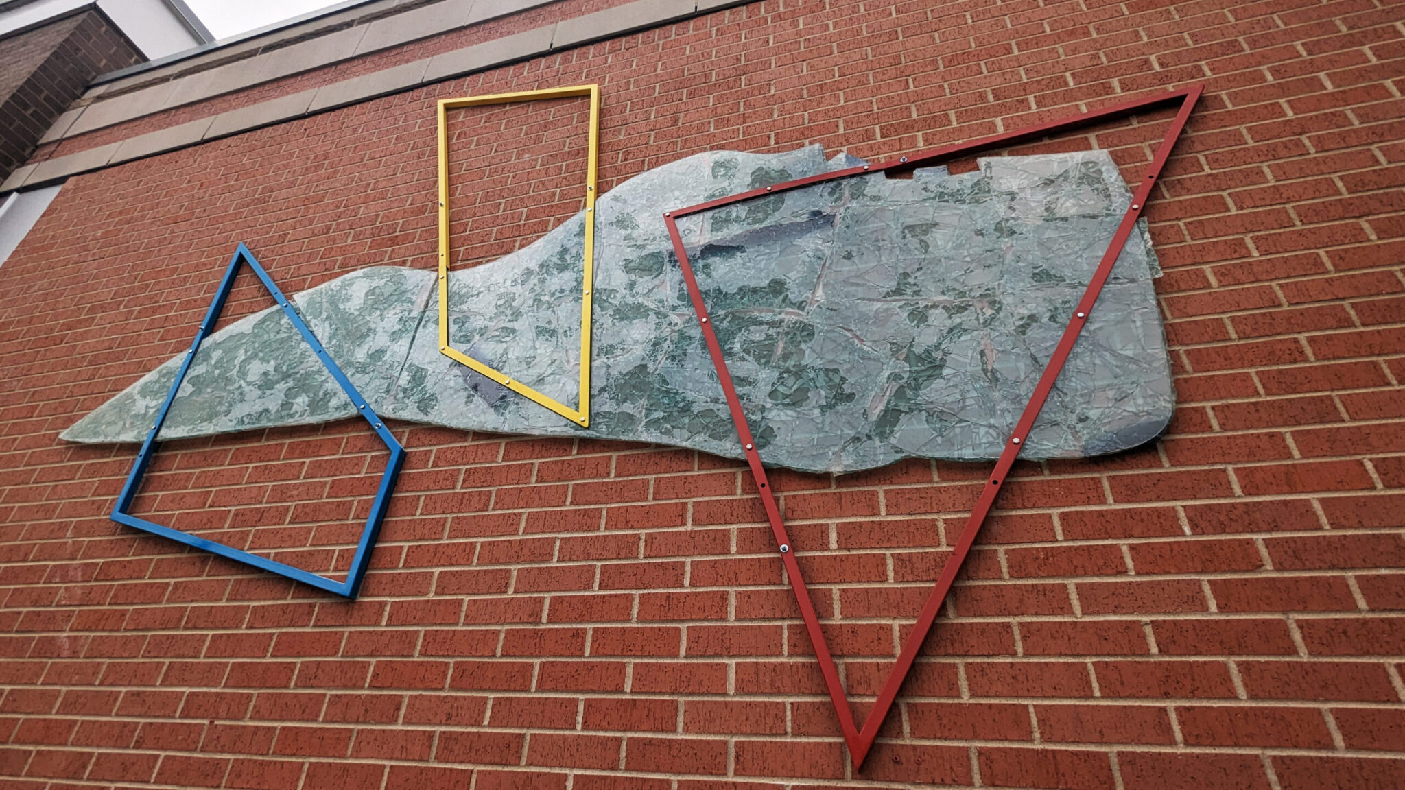 Three metal geometric outlines in blue, yellow and red are bolted against a piece of shattered glass that looks like a map. The shapes represent the redlining of city space. The piece is affixed to a brick wall as part of a series of installations at Chestnut Hill Square.