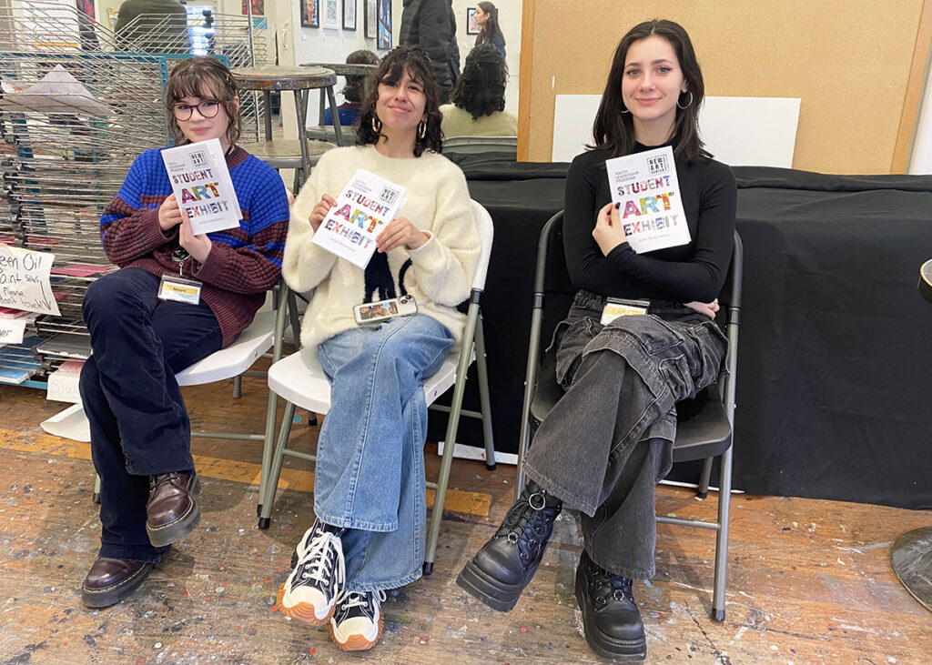 Three Youth Leaders are seated and each are holding the program they made for their teen pop-up exhibition.