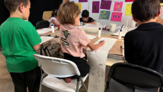 Children engaged in a classroom activity, sitting at tables with paper and scissors, fostering creativity and collaboration.