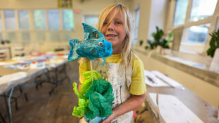 A young girl proudly holds a colorful paper fish, showcasing her creativity and artistic skills in a joyful moment.